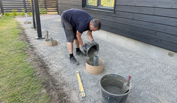 A worker pours concrete into round formwork. A plywood template keeps the steel post bases aligned.