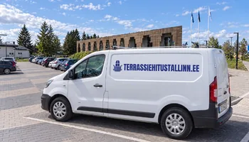 Белый фургон TERRASSIEHITUSTALLINN.EE перед кирпичным фасадом здания волостной управы Сауэ. Во время весеннего ухода в сезоне 2025 года мы перевернули 63 целые, но покоробившиеся доски.