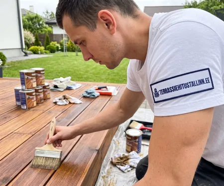 A worker brushes oil onto dark decking beside oil cans and cloths. Used oily cloths go into a water-filled metal container to prevent spontaneous combustion.