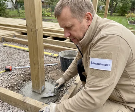 A worker trowels concrete around a post base above gravel. On former marshland in Harju County, screw piles pass through peat to mineral soil.
