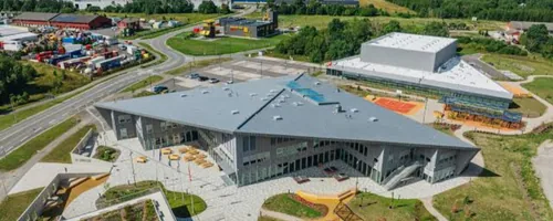 Teravanurgalise halli katusega klaasseintega hoone, selle taga valge kastikujuline saal ja ümber oranžid ning sinised mänguplatsid, arendajalt küsime enne mõõtmist täitepinnase paksuse.