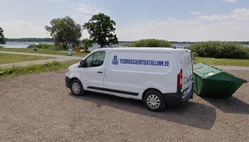 Valge kaubik TERRASSIEHITUSTALLINN.EE kirjaga Maardu järve kruusasel kaldal rohelise prügikonteineri kõrval, taga järv ja lehtpuud, Maardu järve ümbruse kruntidele on ehitatud 11 terrassi.