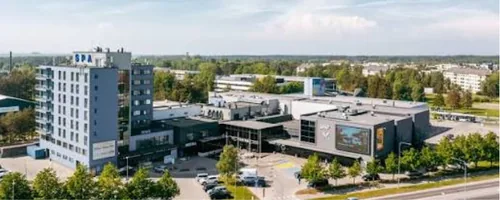 Lõunavalguses Viimsi keskus ülalt: klaasfassaadiga ärihoone suure välisekraaniga, kõrval kõrge hall elamu ja ees puudega ääristatud parkla, rannakrundil hoiame mõõtu lõigatud laudu kuni paigaldamiseni liiva eest presendi all.